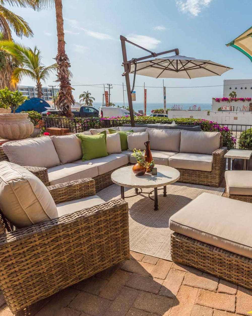 A woven outdoor sectional with cream cushions around a low table, under a cantilever umbrella, palms and the sea beyond