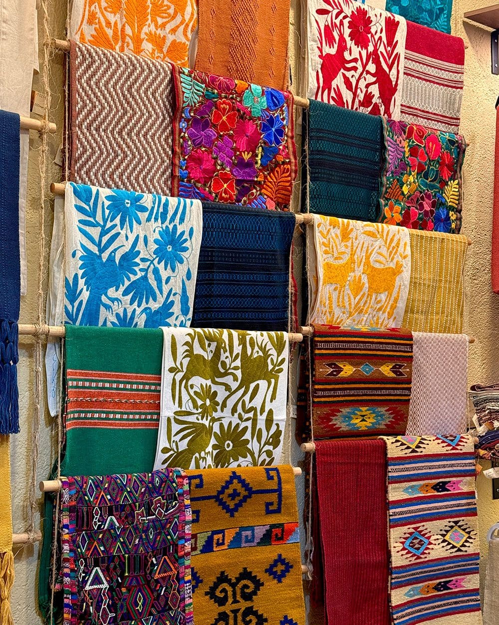 A wall rack of folded textiles, Otomí embroidery, woven runners and Zapotec patterns in many colors