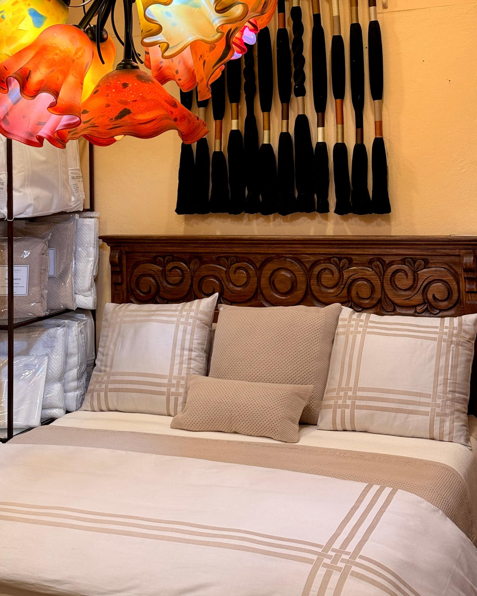 Layered bed linens and woven cushions beneath a carved wooden headboard and blown-glass pendants