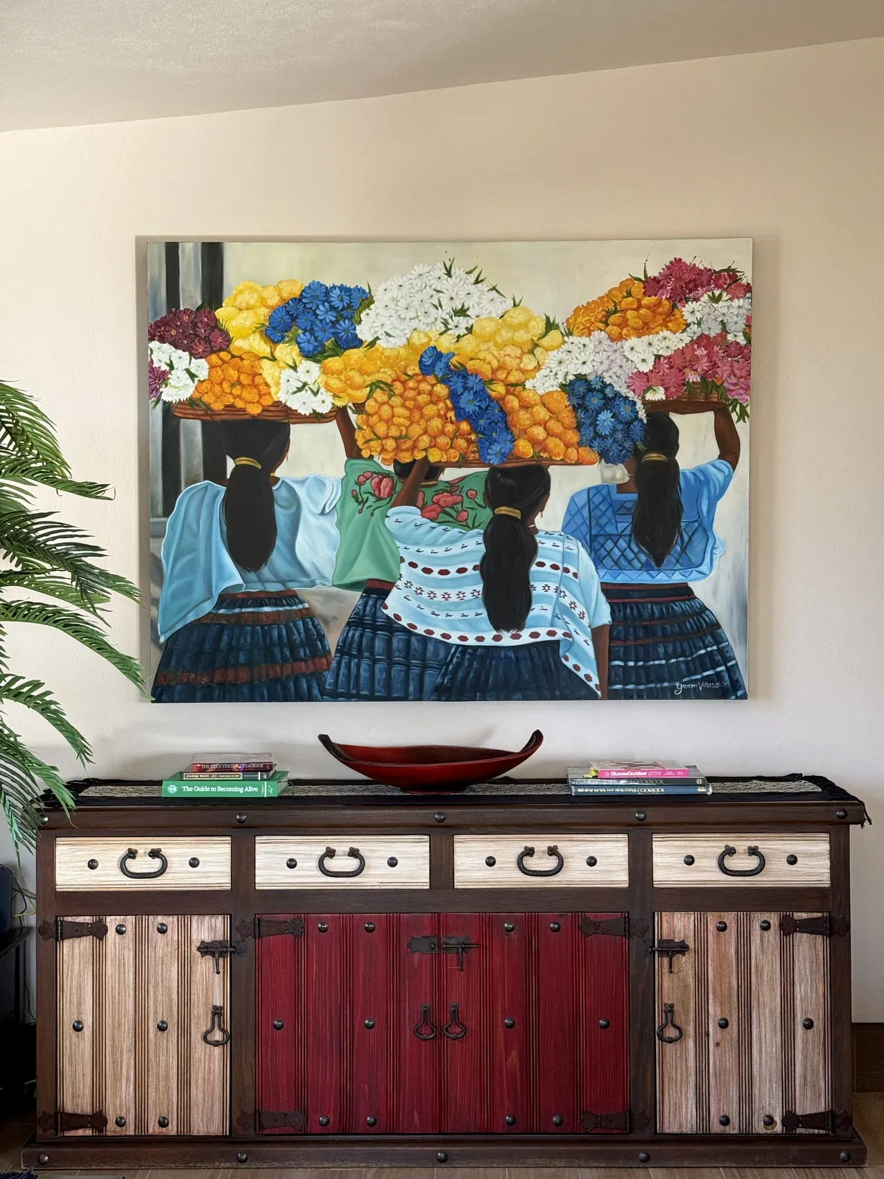 A painting of women carrying baskets of flowers and fruit, hung above a traditional buffet with iron pulls and deep red doors
